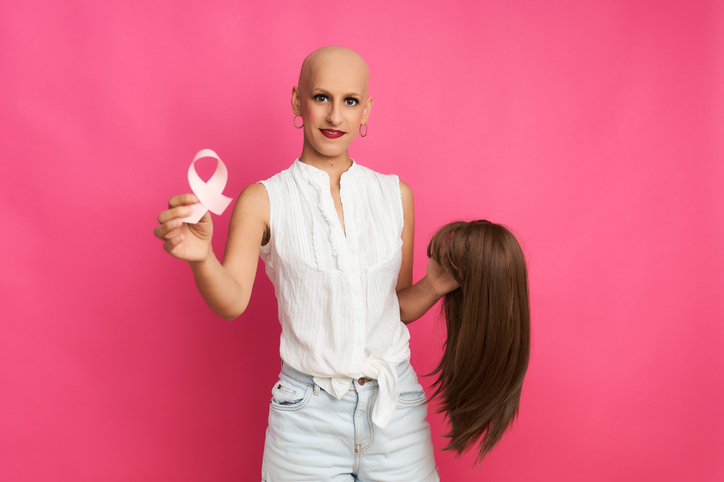 kobieta chora na raka ze wstążką w dłoni i peruką w drugiej ręce na różowym tle